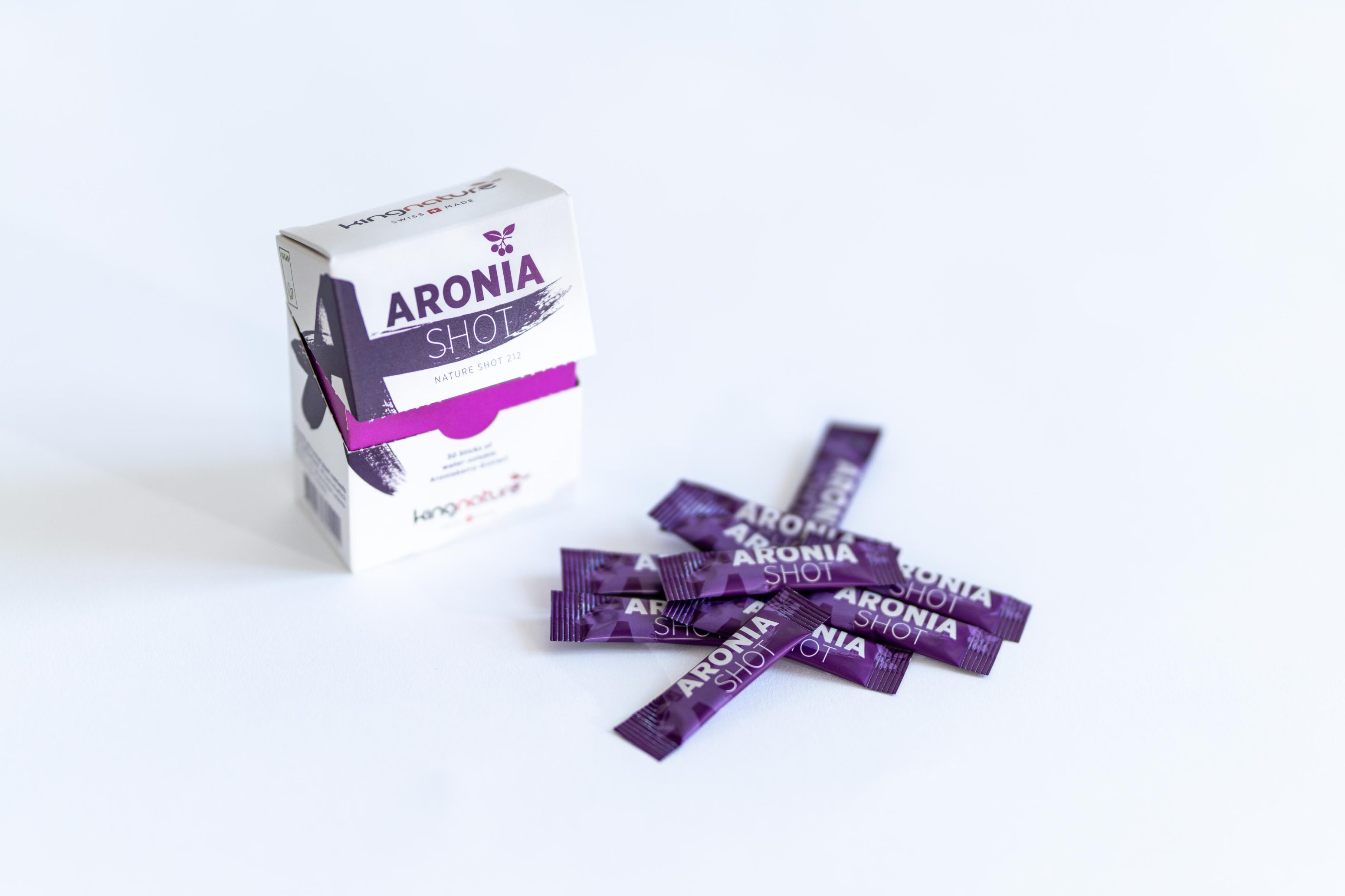 Auf einer weißen Fläche steht eine weiße Schachtel mit der Aufschrift "Aronia Shot", in deren Nähe mehrere lilafarbene Einzelportionspackungen Aronia Shot verstreut sind - ideal für alle, die Aronia Shot kaufen möchten.