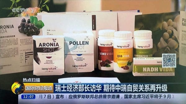 Auf einem Fernsehbildschirm werden Nahrungsergänzungsmittel wie Aronia Shot und Pollen Shot auf einem Tisch mit chinesischen Textbannern, dem CCTV 2-Logo oben links und der Option zum Kauf von Aronia Schokolade angezeigt.
