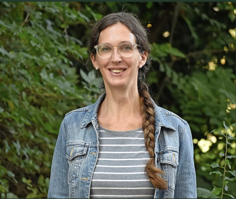 Denyse Yaméogo-Lanter, eine Frau mit langen braunen Haaren zu einem Zopf, Brille, Jeansjacke und grau gestreiftem Hemd, lächelt, während sie draußen vor üppigem grünem Laub steht.