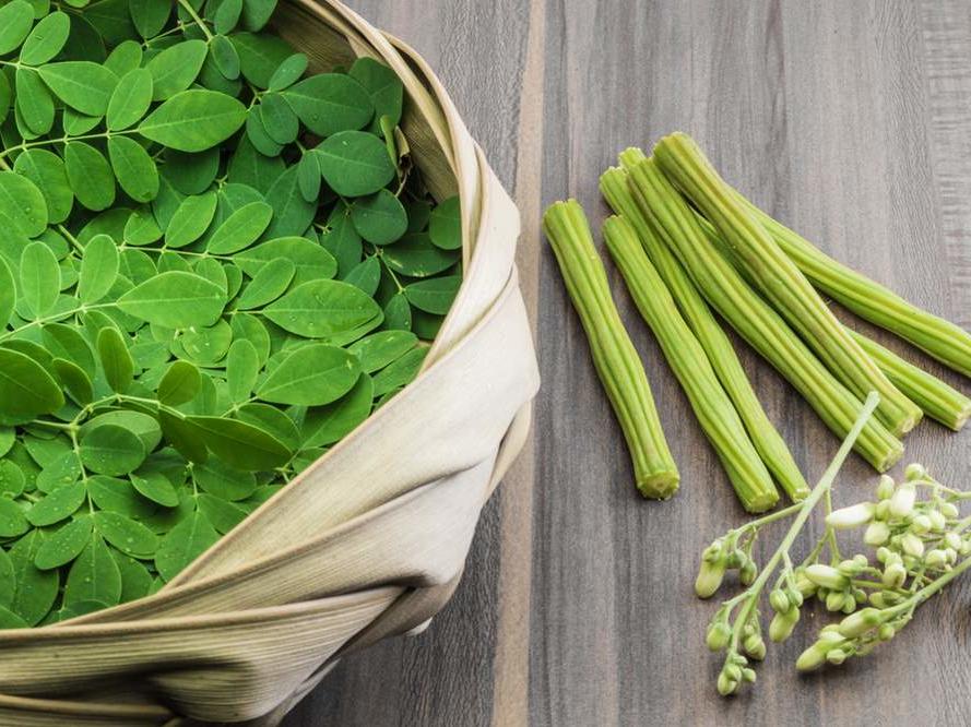 Ein Korb mit frischen grünen Moringa-Blättern, -Schoten und -Blüten steht auf Holz – eine schöne Präsentation für alle, die Bio-Moringa-Blattpulver suchen.