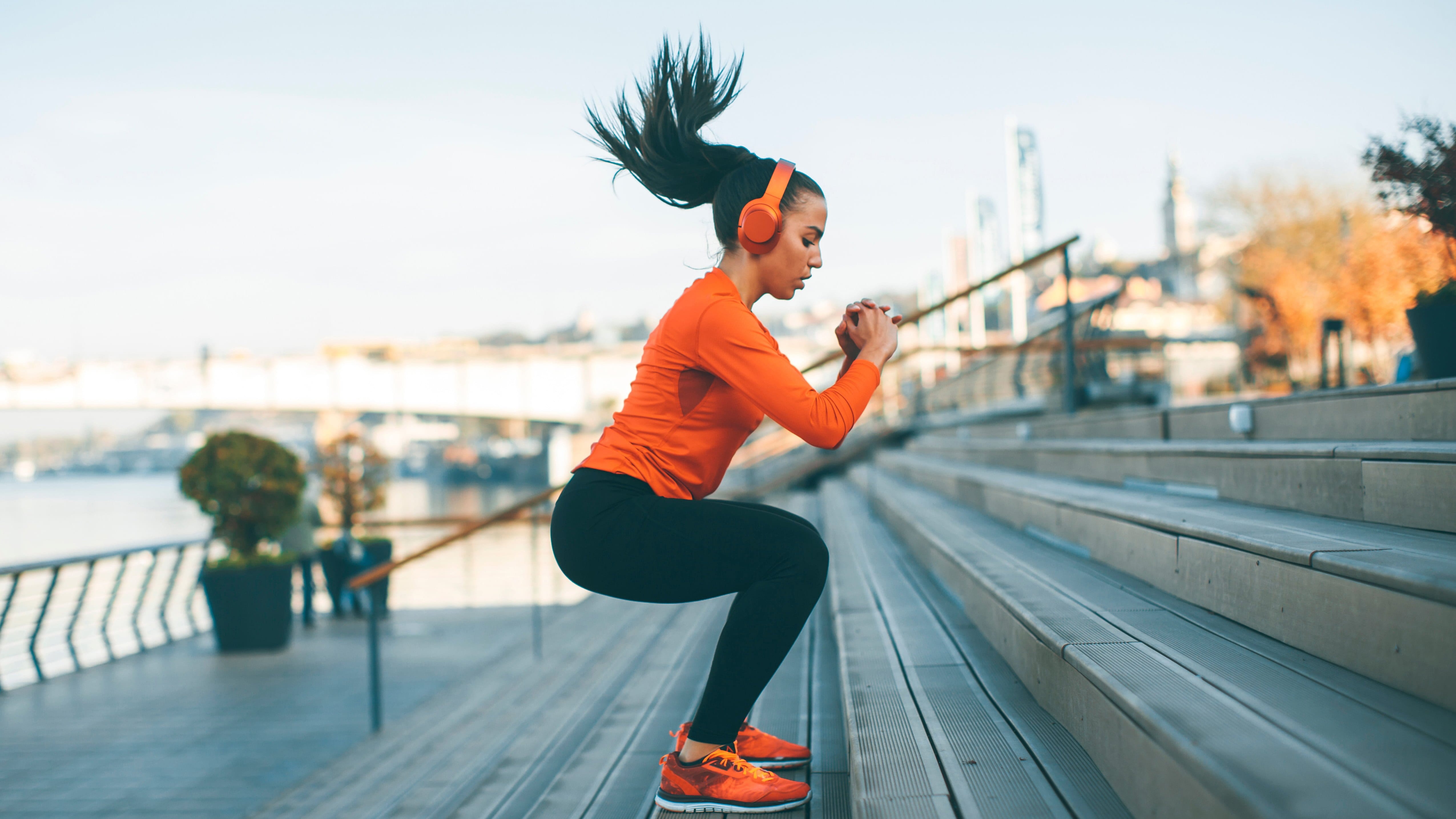 Eine Frau in orangefarbener Sportkleidung und mit Kopfhörern trainiert im Freien und macht einen Hocksprung auf Außentreppen in der Nähe eines Flusses, mit Stadtgebäuden, Bäumen und frischer Luft im Hintergrund.
