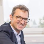 Ein Mann mit kurzen lockigen Haaren und Brille, der einen dunklen Anzug und ein gestreiftes Hemd trägt, lächelt, während er drinnen an einem Fenster sitzt und Selbstvertrauen ausstrahlt.