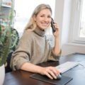 Eine lächelnde Frau mit langen blonden Haaren sitzt an einem Schreibtisch, hält ein Telefon ans Ohr und bedient eine Computermaus. Sie trägt einen grauen Pullover und steht vor einem sonnendurchfluteten Fenster. Eine grüne Pflanze sorgt für Frische am Arbeitsplatz.