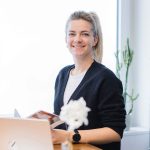 Eine Frau mit blondem Haar und Pferdeschwanz sitzt lächelnd an einem Schreibtisch am Fenster. Sie trägt eine schwarze Strickjacke, ein weißes Hemd, eine Uhr und hält eine Zeitschrift in der Hand. Ein Laptop, eine Blume, ein Kaktus und Sonnenlicht, das durch das Fenster fällt, runden die Szene ab.