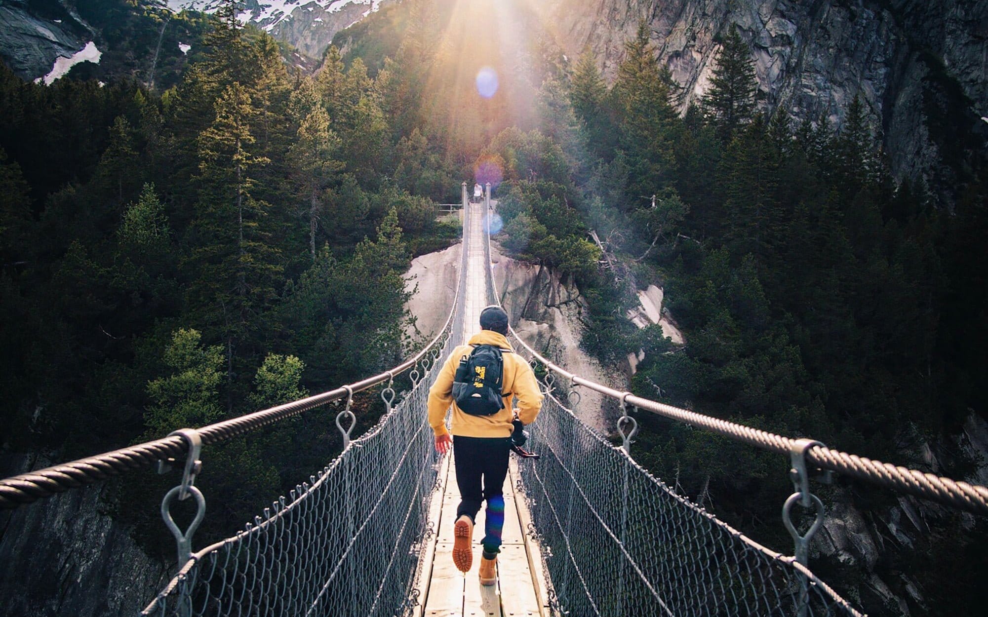 Eine Person in einer gelben Jacke und einem Rucksack geht über eine Hängebrücke, die von dichtem Wald und Bergen umgeben ist, während Sonnenlicht durch die Bäume fällt.